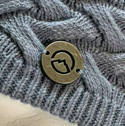 Close up of Domex medallion on  Dusty blue skullcap beanie with accent woven cable knit band 